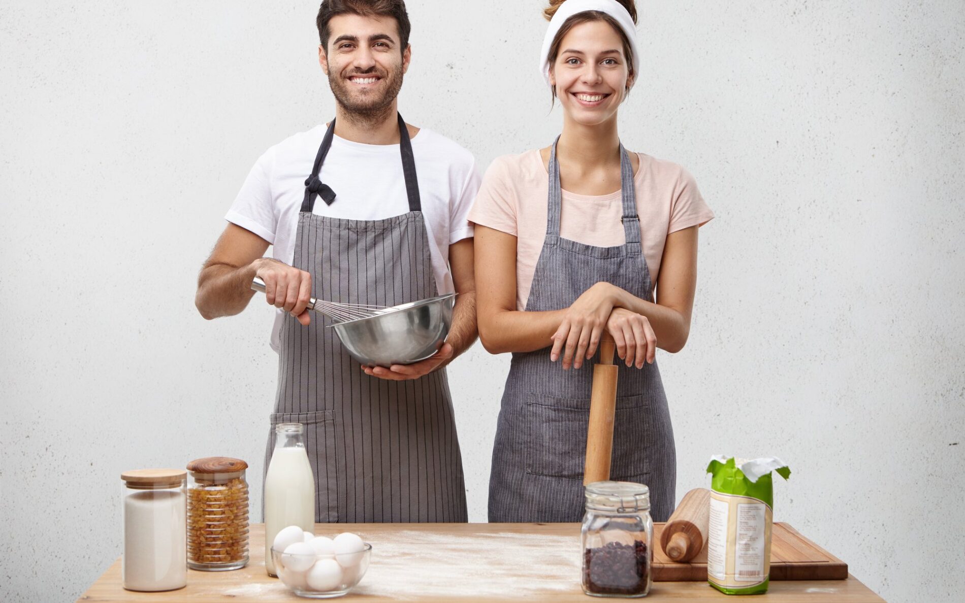 Young family couple standing on kitchen, cooking dinner together, surrounded with different products and tablewear, having happy expressions. Joyful man and woman preparing pancake for breakfast